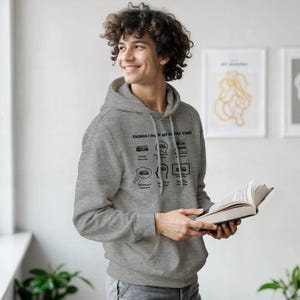 Peut inclure: Un sweat à capuche gris avec un graphique indiquant "THINGS I DO IN MY SPARE TIME" avec des illustrations d'un bus, de bulles de pensée et d'un ordinateur portable. Le sweat à capuche a une capuche à cordon et une poche avant.