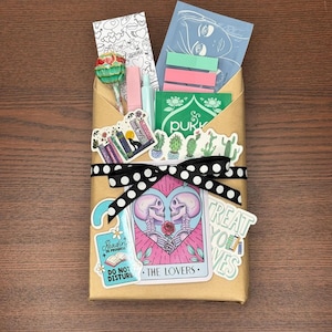 May include: A brown paper-wrapped gift box tied with a black and white polka dot ribbon. The box is decorated with stickers, including a tarot card design with skeletons and the words "The Lovers." The box contains various items, including a lollipop and stickers.