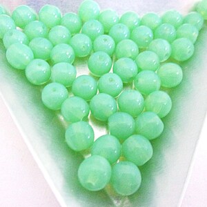 May include: A collection of round, light green beads arranged in a triangular shape. The beads have a smooth, glossy surface and are displayed on a light green, triangular dish. These beads are suitable for jewelry making and crafting.