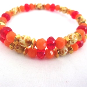 May include: A multi-strand wrap bracelet featuring faceted beads in vibrant shades of red and orange, interspersed with round, speckled beige beads and small, metallic gold-toned spacers arranged in a repeating pattern.