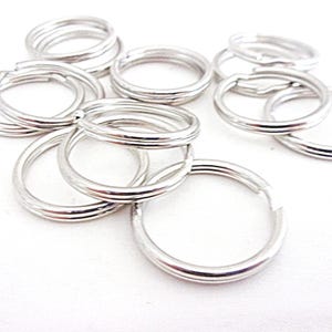 May include: A collection of silver-colored split key rings, scattered on a white surface. The key rings are round, with a split in the ring for attaching keys or other items. The rings are made of a shiny metal.