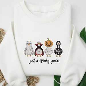 May include: White sweatshirt with a Halloween-themed design. The design features four cartoon geese dressed as a ghost, vampire, pumpkin, and skeleton. The text "just a spooky goose" is below the geese.