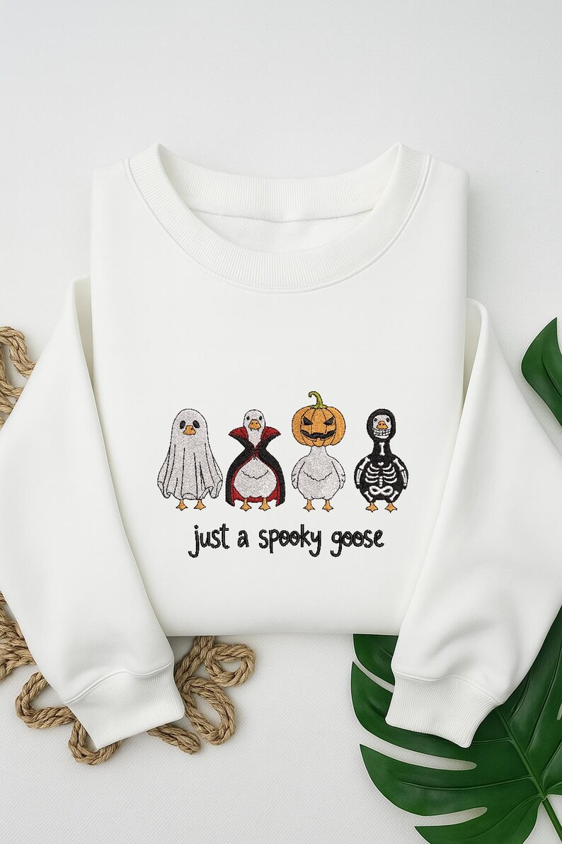 May include: White sweatshirt with a Halloween-themed design. The design features four cartoon geese dressed as a ghost, vampire, pumpkin, and skeleton. The text "just a spooky goose" is below the geese.