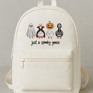 May include: Off-white backpack with a front zippered pocket and adjustable straps. The front of the bag features embroidered cartoon geese dressed as a ghost, vampire, jack-o'-lantern, and skeleton. Text below the geese reads "just a spooky goose."