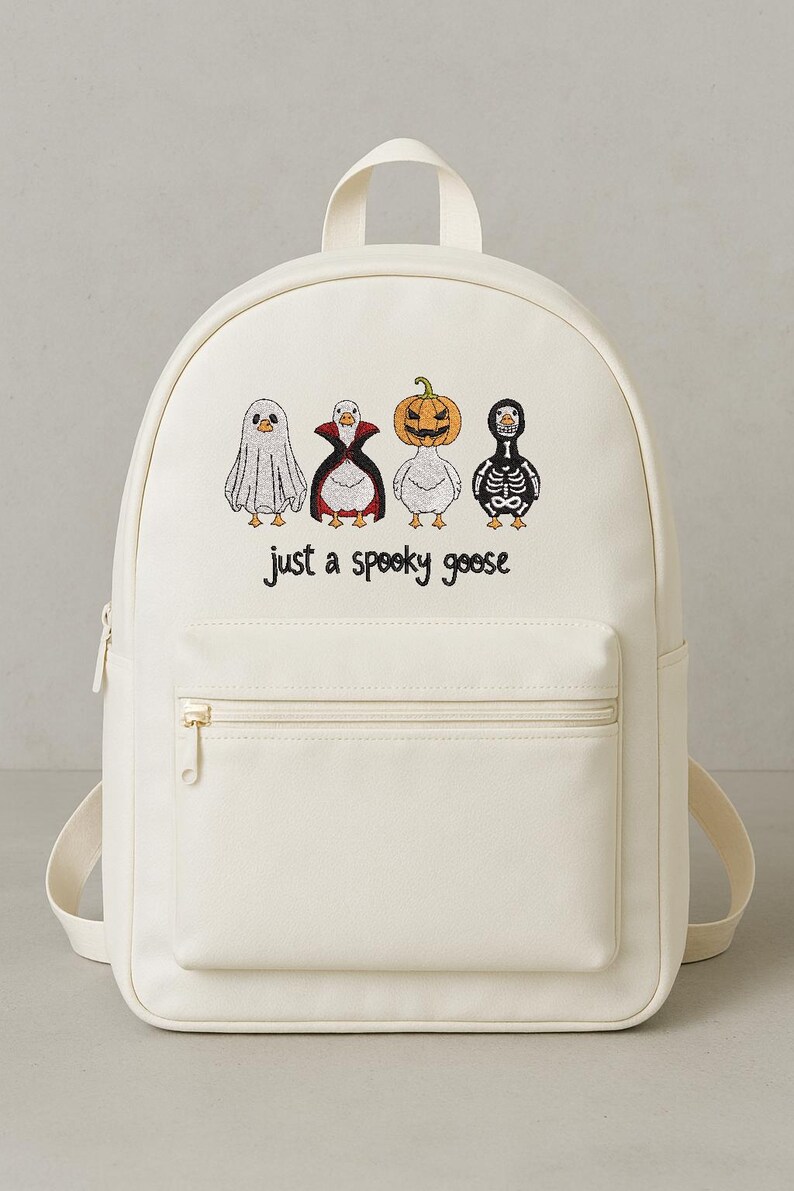 May include: Off-white backpack with a front zippered pocket and adjustable straps. The front of the bag features embroidered cartoon geese dressed as a ghost, vampire, jack-o'-lantern, and skeleton. Text below the geese reads "just a spooky goose."