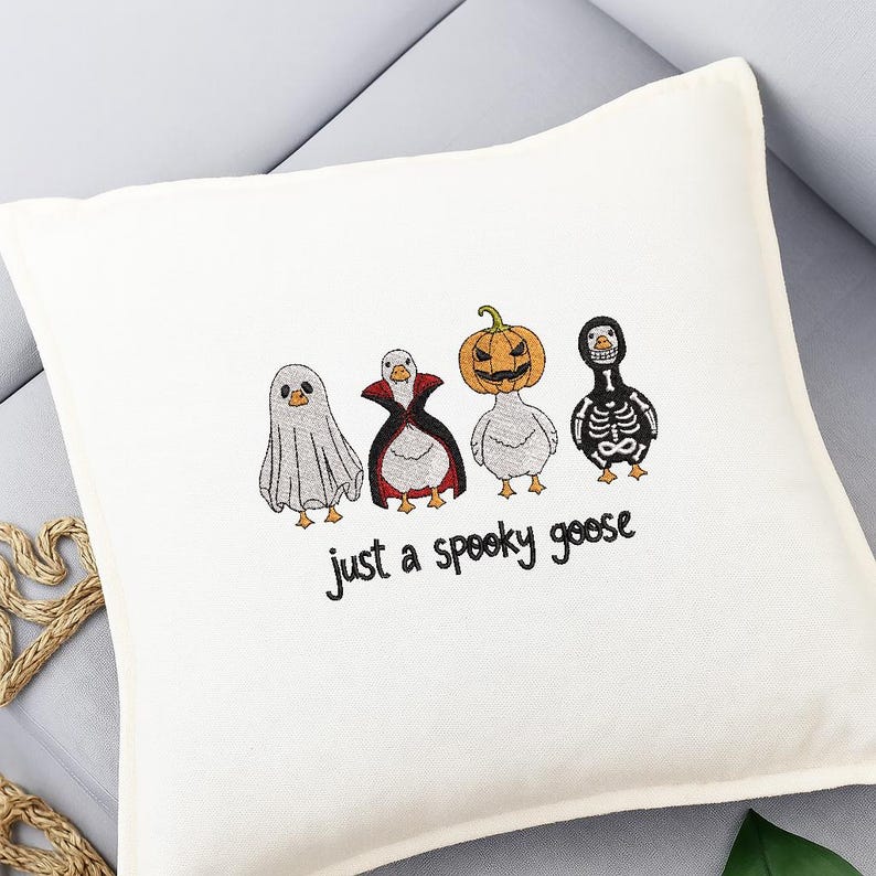 May include: Off-white square pillow with embroidered Halloween geese. The geese are dressed as a ghost, vampire, jack-o'-lantern, and skeleton. The text "just a spooky goose" is embroidered below the geese.