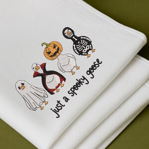 May include: White folded cloth with embroidered Halloween-themed geese. The geese are dressed as a ghost, vampire, jack-o'-lantern, and skeleton. The text "just a spooky goose" is embroidered in black.