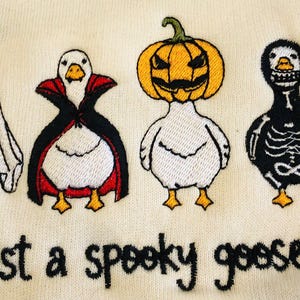 May include: Embroidered design featuring four geese dressed in Halloween costumes: a ghost, a vampire, a jack-o'-lantern, and a skeleton. The text "just a spooky goose" is embroidered below the geese. The geese are white with yellow beaks and feet.