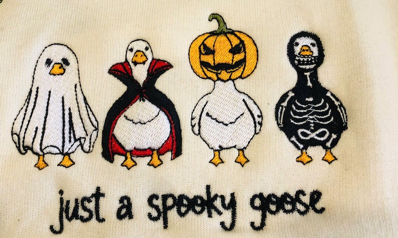 May include: Embroidered design featuring four geese dressed in Halloween costumes: a ghost, a vampire, a jack-o'-lantern, and a skeleton. The text "just a spooky goose" is embroidered below the geese. The geese are white with yellow beaks and feet.