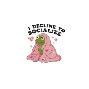 May include: An embroidered design of a green frog character wrapped in a pink blanket decorated with hearts and a flower. The text "I DECLINE TO SOCIALIZE" is above the frog, with star accents.