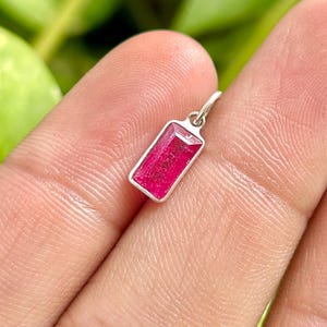 Könnte beinhalten: Ein kleiner, rechteckiger Anhänger mit einem leuchtend rosa Edelstein in einem silbernen Rahmen. Der Edelstein hat eine rechteckige Form. Der Anhänger hängt an einer kleinen Silberschlaufe.