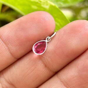 May include: A small, teardrop-shaped pendant with a vibrant pink gemstone set in a silver bezel. The pendant is suspended from a small silver loop, perfect for a necklace or bracelet. The gemstone is a rich, saturated color.