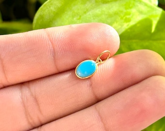 Peut inclure: Un petit pendentif ovale avec une pierre turquoise vive sertie dans un métal doré. Le pendentif a un design simple avec une boucle en haut pour une chaîne. L'arrière-plan est flou, avec des feuilles vertes.