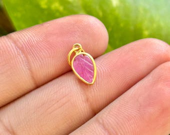 May include: A small, leaf-shaped pendant with a vibrant pink gemstone set in a gold-colored frame. The pendant is held against a blurred background of green leaves and skin.