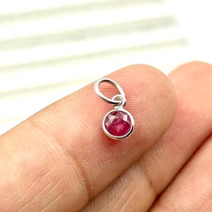 May include: A small, round, ruby gemstone pendant with a faceted surface. The ruby is set in a silver-colored metal bezel and has a small loop at the top for attaching to a chain. The pendant is held up by a finger.