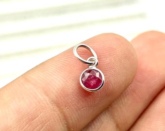 May include: A small, round, ruby gemstone pendant with a faceted surface. The ruby is set in a silver-colored metal bezel and has a small loop at the top for attaching to a chain. The pendant is held up by a finger.