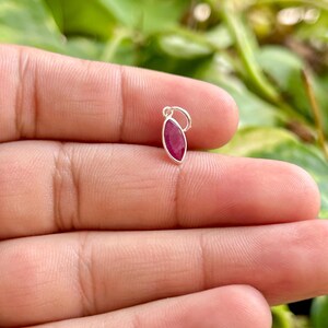 May include: A small, oval-shaped ruby pendant with a silver bail. The ruby is a deep red color and is set in a polished silver setting. The pendant is held in a hand against a blurred green background.