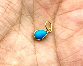 May include: A gold pendant with a teardrop-shaped, bright turquoise stone. The pendant has a simple, elegant design, with a gold frame around the stone and a small loop for a chain. The pendant is small and delicate.