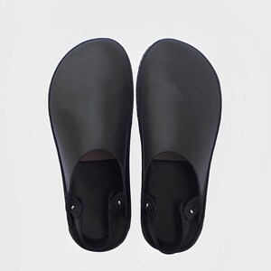Könnte beinhalten: Ein Paar schwarze Lederclogs mit rundem Zehenbereich. Die Clogs haben einen Fersenriemen und sind vor einem weißen Hintergrund platziert. Die Schuhe scheinen neu zu sein.
