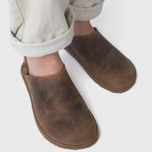 Może przedstawiać: Para brązowych skórzanych chodaków z postarzanym wykończeniem. Chodaki mają zaokrąglony nosek i niskoprofilową podeszwę. Buty są noszone ze spodniami w kolorze kremowym.