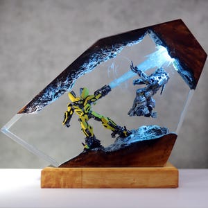 May include: A resin sculpture featuring two action figures engaged in battle, encased in a clear, angled block. One figure is yellow and black, the other is silver. The scene is illuminated with blue light, set on a wooden base.