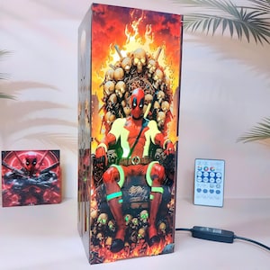 Può includere: Lampada rettangolare con un'illustrazione a fumetti di Deadpool seduto su un trono di teschi, con uno sfondo infuocato. Sono presenti anche un telecomando e una piccola immagine quadrata di Deadpool. La lampada è collegata.
