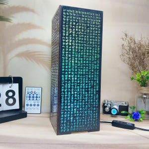 May include: A rectangular, dark-colored lamp with a matrix of green illuminated characters. The lamp is on a wooden surface, next to a calendar, remote, and a vintage camera. The lamp has a power cord.