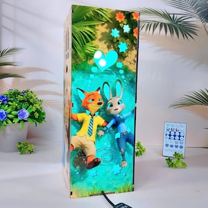 Puede incluir: Una caja de luz de madera con los personajes Nick Wilde y Judy Hopps de Zootopia. La caja tiene una colorida ilustración de los personajes en una escena acuática con corazones y estrellas. Se ve un mando a distancia.