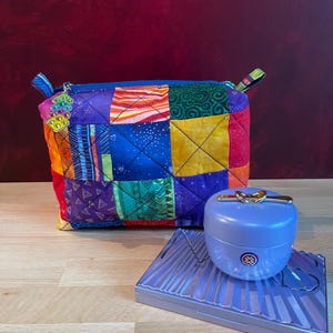 May include: A colorful patchwork zippered pouch with a blue zipper and small charm. A light blue cosmetic container with a gold-colored bow detail sits on a matching rectangular compact.