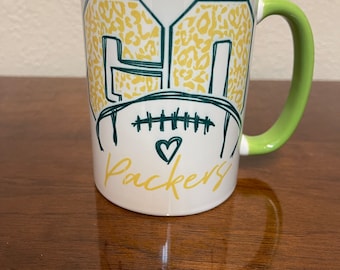 May include: White ceramic mug with a green handle. The mug has a green and yellow design with the number "GO" and a football with a heart above the word "Packers".
