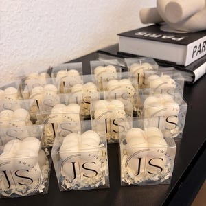 Peut inclure: Plusieurs bougies blanches en forme de nuage, présentées dans des boîtes transparentes. Chaque bougie porte les initiales "JS" et les noms "JULIA" et "SAMIR". Elles sont disposées sur une surface noire.