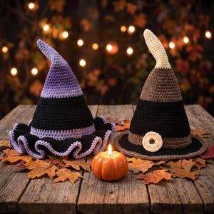 May include: Two crocheted witch hats. The left hat features a purple-to-black ombre design with a ruffled brim. The right hat is brown and black with a cream-colored tip and a small circular accent. A small orange pumpkin candle sits between them.