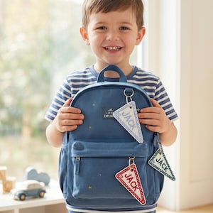 May include: A blue backpack featuring a small rectangular patch with the text LITTLE EXPLORER and scattered gold star patterns. Three triangular pennant-shaped keychains are attached to the bag, each embroidered with the name JACK.