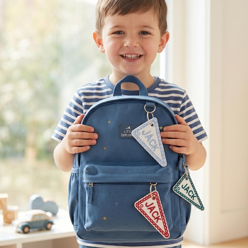 May include: A blue backpack featuring a small rectangular patch with the text LITTLE EXPLORER and scattered gold star patterns. Three triangular pennant-shaped keychains are attached to the bag, each embroidered with the name JACK.