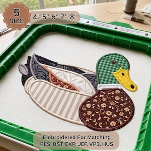 May include: An embroidered mallard duck design featuring a green checkered head, a yellow beak with black dots, and a body composed of layered fabric patterns including brown floral, beige stripes, and dark gray leaf motifs, set in a green embroidery hoop.