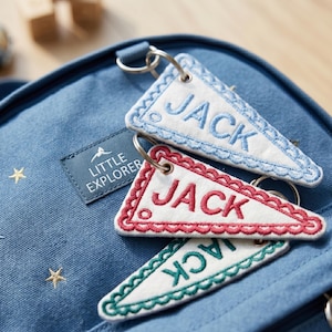 May include: Three triangular white fabric keychains with scalloped edges and embroidered text reading JACK in blue, red, and green. The keychains are attached to a blue canvas bag featuring gold star embroidery and a patch labeled LITTLE EXPLORER.