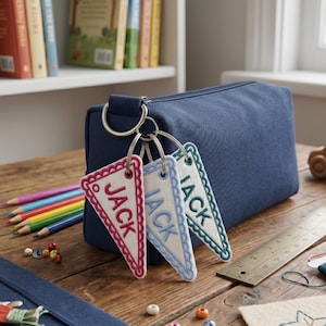 May include: A navy blue canvas pencil case with three triangular felt pennant keychains attached to a metal ring. Each white pennant features a scalloped edge and the name JACK embroidered in red, blue, or green thread respectively.