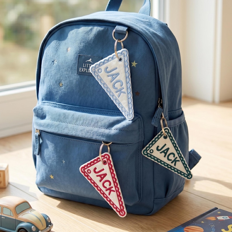 May include: A blue backpack featuring a small rectangular patch with the text LITTLE EXPLORER and scattered star embroidery. Three triangular pennant-shaped keychains are attached to the zippers, each embroidered with the name JACK.