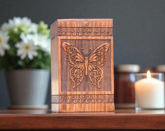 Puede incluir: Una caja de madera con un diseño de mariposa tallada. La mariposa está centrada en el panel frontal, rodeada de tallas florales. La caja tiene patrones florales decorativos en la parte superior e inferior. La madera tiene un tono cálido y natural.