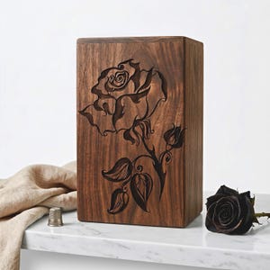 Op de afbeelding: Een rechthoekige houten kist met een gebeeldhouwd rozenontwerp. De kist is gemaakt van donkerbruin hout en heeft een gedetailleerde bloemengravure van een roos met bladeren en knoppen. Een zwarte roos ligt naast de kist.