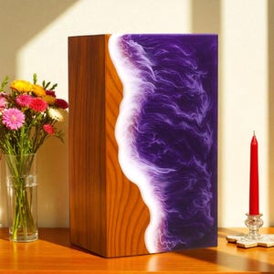 May include: A decorative wooden box with a unique design. One side is natural wood, the other has a purple resin with a white, wave-like pattern. A red candle and a vase of flowers are also visible.