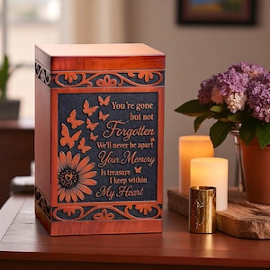 Può includere: Urna in legno marrone con pannello centrale blu scuro con girasole, farfalle incise e la frase "You're gone but not Forgotten". L'urna presenta intagli decorativi.