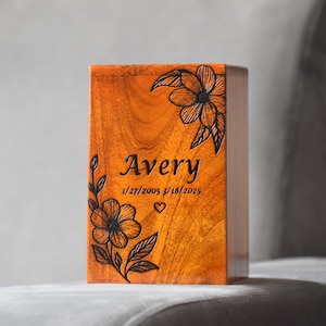 Può includere: Scatola commemorativa in legno con finitura arancione caldo. La scatola è decorata con disegni floreali neri e il nome "Avery" insieme alle date. È incluso anche un simbolo a forma di cuore.
