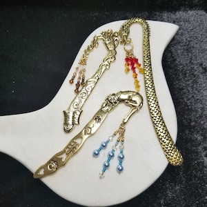Könnte beinhalten: Drei goldene Lesezeichen aus Metall mit dekorativen Designs und baumelnden Perlen. Die Lesezeichen befinden sich auf einem weißen, vogelartigen Teller. Ein Lesezeichen ist ein Drache, eines ist eine Tasse und eines ist ein Fisch.