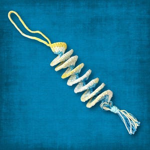 Könnte beinhalten: Handgefertigter Spiralanhänger aus Häkelarbeit in Weiß, Gelb und Blau. Das Ornament besteht aus einer Reihe kreisförmiger Scheiben, die durch eine gehäkelte Kordel verbunden sind, mit einer Quaste unten und einer Schlaufe oben zum Aufhängen.