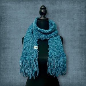 May include: A teal-colored knitted scarf with fringe detail. The scarf is displayed on a black mannequin. A small white tag is attached to the scarf. The scarf is made of a soft, textured yarn.