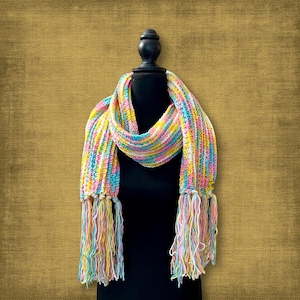 May include: A pastel-coloured, handmade scarf displayed on a black mannequin. The scarf has a striped pattern in pink, yellow, blue, and white. The ends of the scarf are decorated with long, matching fringe. The scarf is a cosy accessory.