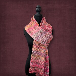 May include: A colorful, hand-knitted scarf displayed on a black mannequin. The scarf features a textured pattern and a blend of pink, orange, and gray hues. The scarf is wrapped around the mannequin's neck and drapes down.