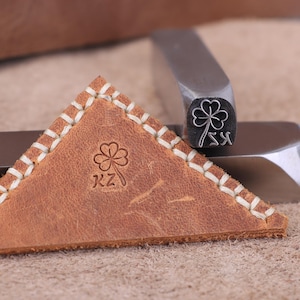 Op de afbeelding: Een bruin leren item met een gestikte rand en een klaverontwerp. Een metalen stempelgereedschap met een klaverontwerp staat naast het leren item. Het leren item heeft de letters "KZ" eronder gestempeld.
