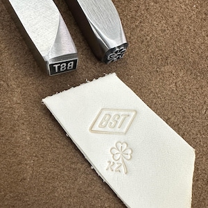 May include: Close-up of leather stamping tools and stamped leather. Two metal stamps are visible, one with the text "T89" and the other with a floral design. A white leather piece displays stamped text and a clover design. The leather is on a brown surface.